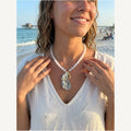 Hera necklace of pale white jade rounds with a lavender-glazed oyster shell and gold mermaid charm, worn on the beach