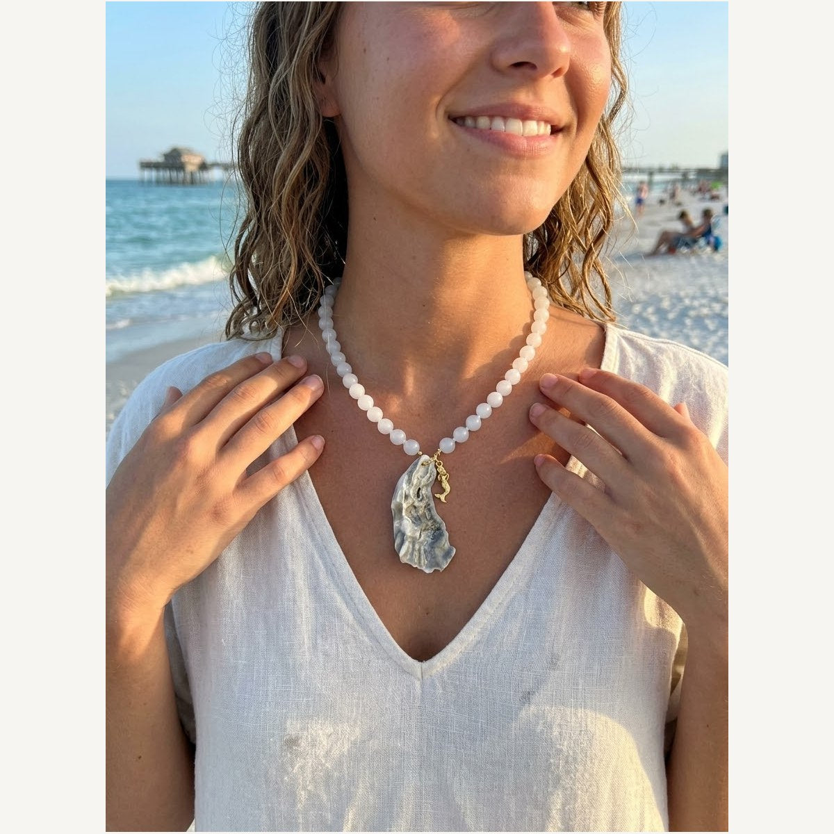 Hera necklace of pale white jade rounds with a lavender-glazed oyster shell and gold mermaid charm, worn on the beach